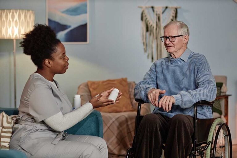 Elderly resident discussing health questions with a nurse at a table, with medication organizer and a notepad present.